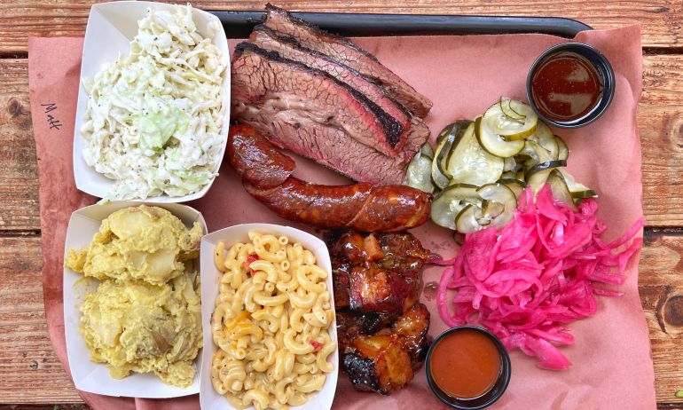 a tray of barbecued meat, sauces, sides of pickle and cucumber, and coleslaw, mashed potatoes and macaroni and cheese