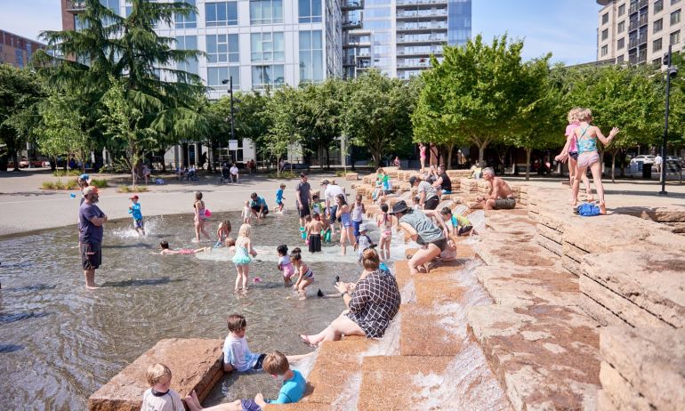 many children and a few adults play in the fountain at Jamison Square on a bright summer day