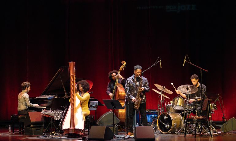 A band of five Jazz musicians performs on stage in front of a red curtain.
