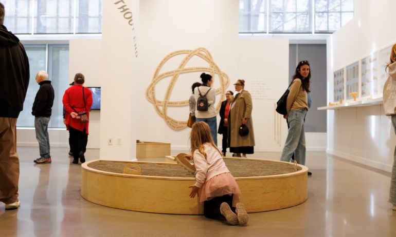 several people browse hung art on the walls of a museum while a child plays in a central sand box