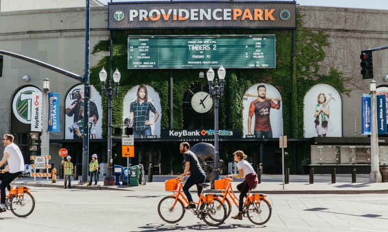 two cyclists on Biketown bikes pedal past Providence Park exterior