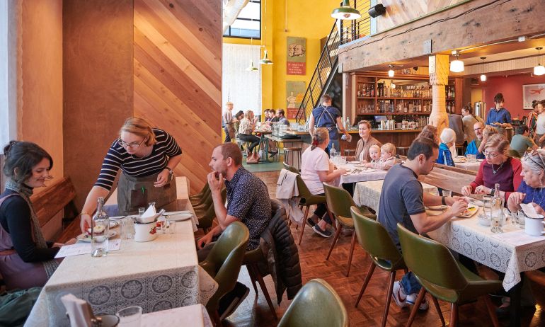 tables full of diners enjoy a meal in a lively, warmly lit restaurant with a bar in the background