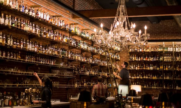 several bartenders peruse shelves housing a vast collection of whiskey bottles