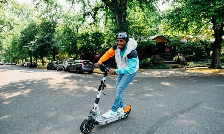 a smiling man wearing a helmet rides a Biketown scooter on a leafy city street