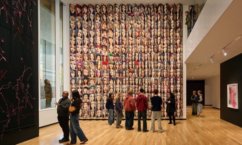 People look at art, including a large, floor-to-ceiling installation featuring many faces, from several different angles: from outside a window, from a ground floor view, and looking over a balcony.