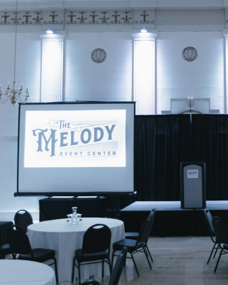 tables and chairs set for an event with two screens and a podium in front at Melody Event Space