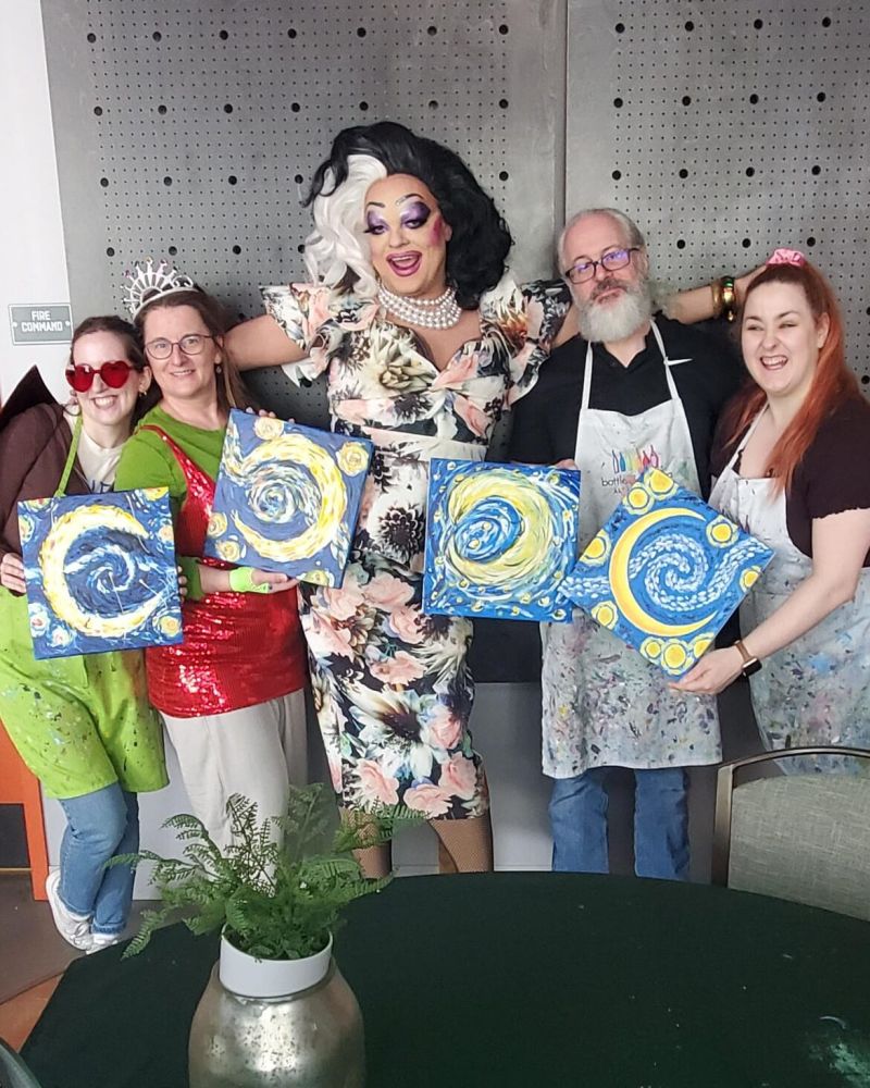 A group of smiling people wearing paint-covered aprons pose with their paintings, standing next to a smiling drag queen