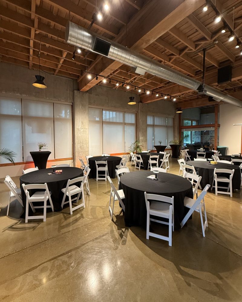 An industrial event venue with round tables draped in black tablecloths and white chairs.