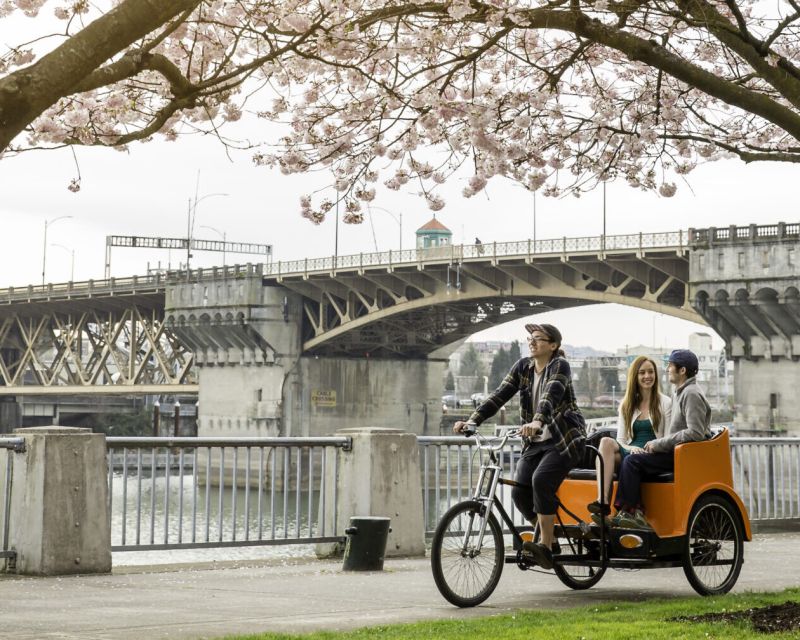 orange pedicab rides through park under backlit cherry blossoms and a view of the Burnside Bridge