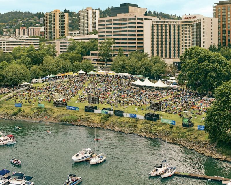 an arial view of a large crowd of people, stages and tented vendors in a park on the waterfront of the Willamette River with several boats floating in the water and a skyline view of downtown Portland in the background
