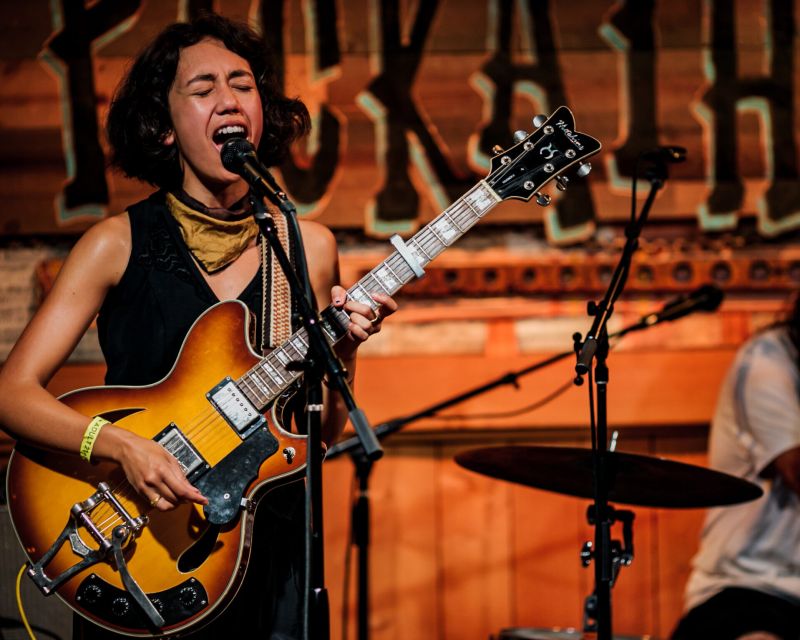 person singing at a microphone with a guitar, a drummer is in the background