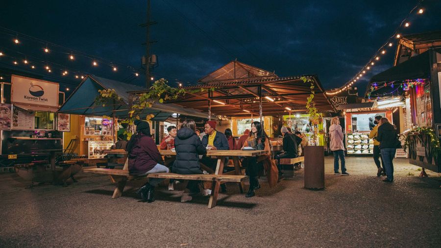 people seated at picnic tables talk and eat with food carts in the background