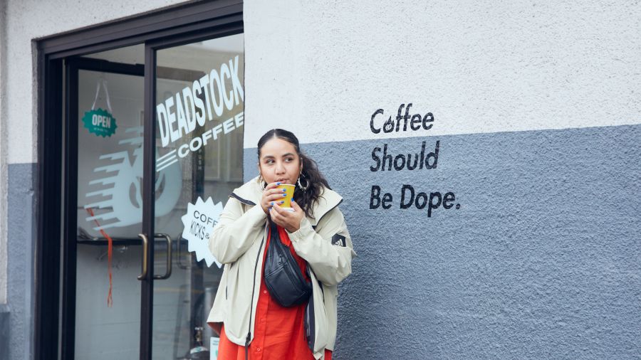 person stand outside of a store front with a coffee there is a mural on the building that says Coffee Should Be Dope