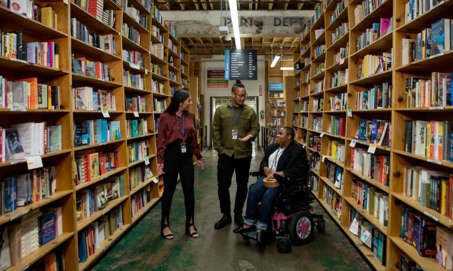 three people talk in a bookstore aisle; two are standing, one is in a wheelchair