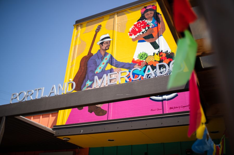 A sign reading "Portland Mercado" in front of a brightly painted mural showing a Latin American dancer and musician