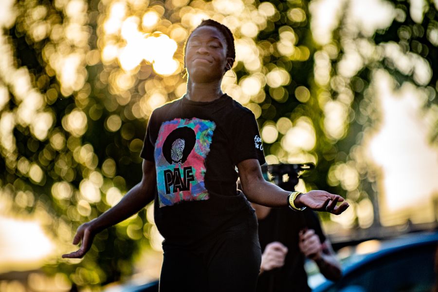 woman holds hands and arms out in dance performance in a park, sun filters through the leaves of the tree behind her