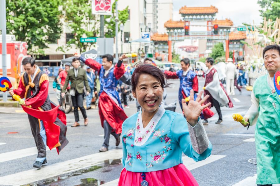 woman with big smile waves in a parade with colorful costumes on all the folks in the frame Chinese gate in background