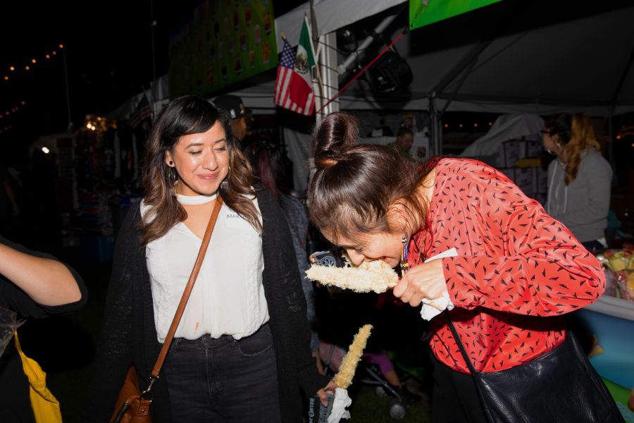 two people standing next to a food cart, one eating an ear of corn