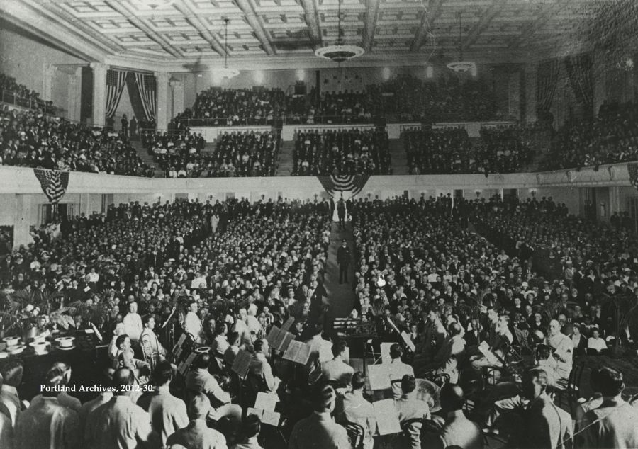 Historic black and white photo of people gathered in a packed auditorium around an orchestra.