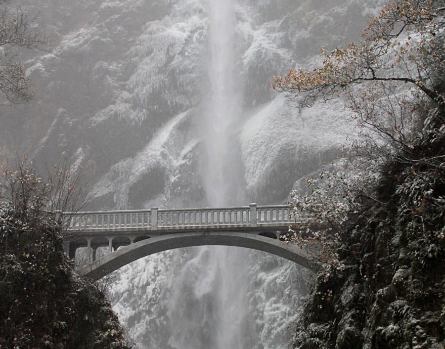 a towering waterfall behind a stone bridge surrounded by snow and ice