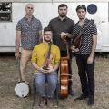 All four member of Invoke casually dressed posing with their instruments outside in front of an old Airstream trailer.