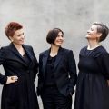 The three women of Trio Mediaeval dressed in black clothes looking at each other smiling.