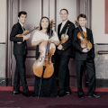 Members of Calidore Quartet smiling, dressed in formal wear in black in white, holding their instruments standing in front of double doors.