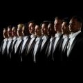 Members of Chanticleer dressed in tuxedos standing in a line with shadows casted on their faces.