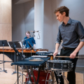 Photo of a percussion performer playing a snare drum, with a marimba player in the background.