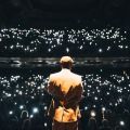 the back of a person on a stage facing an audience full pf people shining lights at the performer
