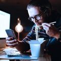 a person in front of a computer screen eating noodles and looking at a phone