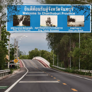 ถนนลาดผ่านเนินใต้ป้ายสีน้ำเงินที่เขียนว่ายินดีต้อนรับสู่จังหวัดจันทบุรี มีต้นไม้เรียงอยู่สองข้างทาง