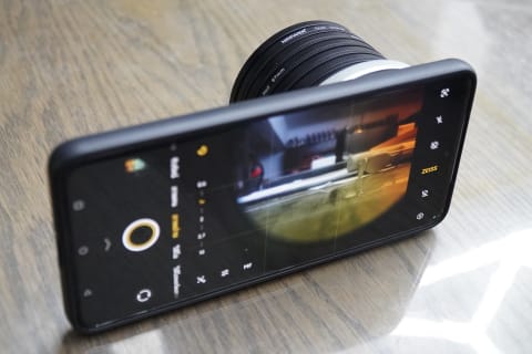 Smartphone with a camera lens attachment positioned on a wooden table, displaying a camera interface on the screen.