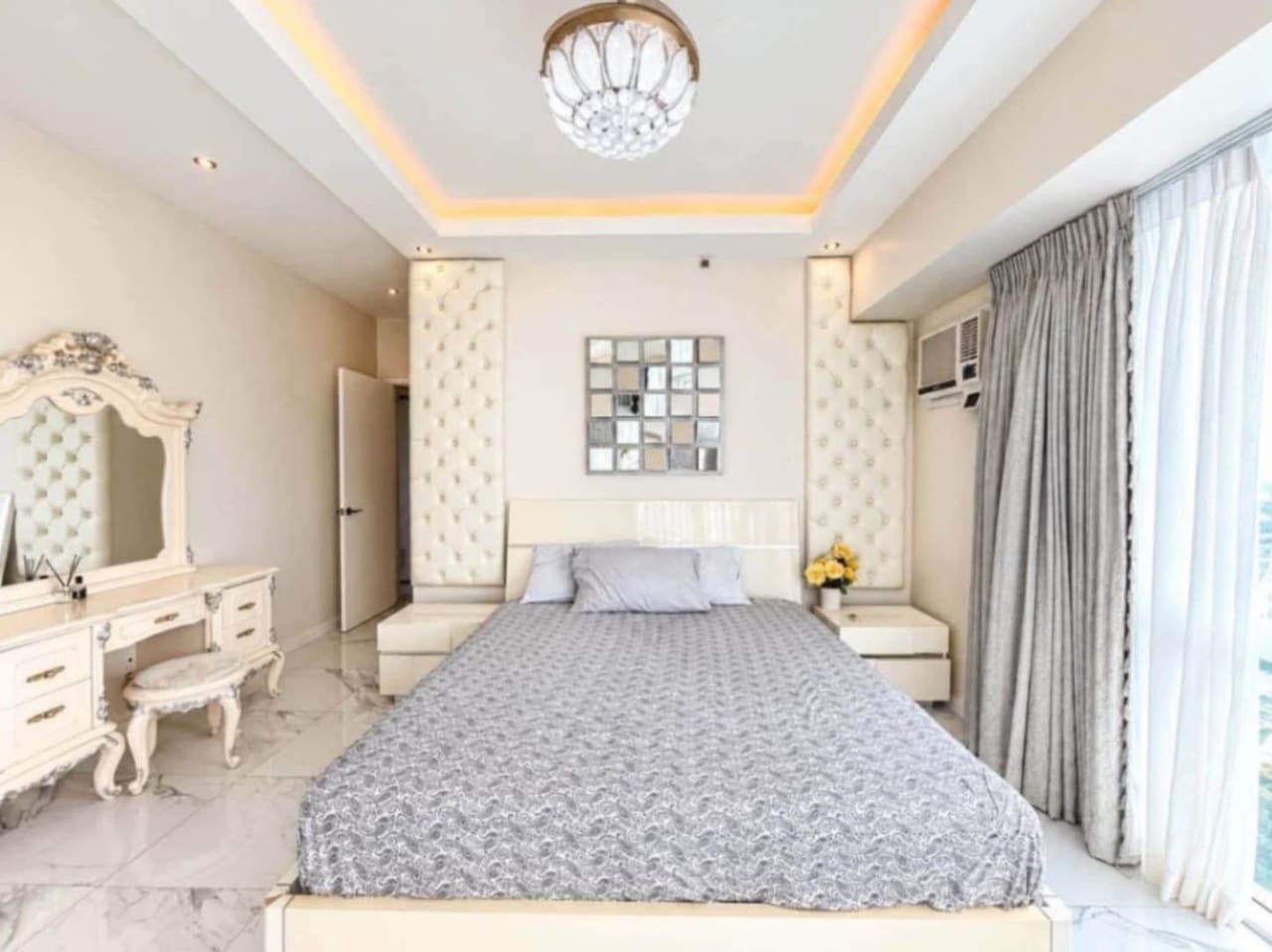 Master bedroom with tufted headboard panels, vanity dresser, crystal chandelier and tray ceiling lighting