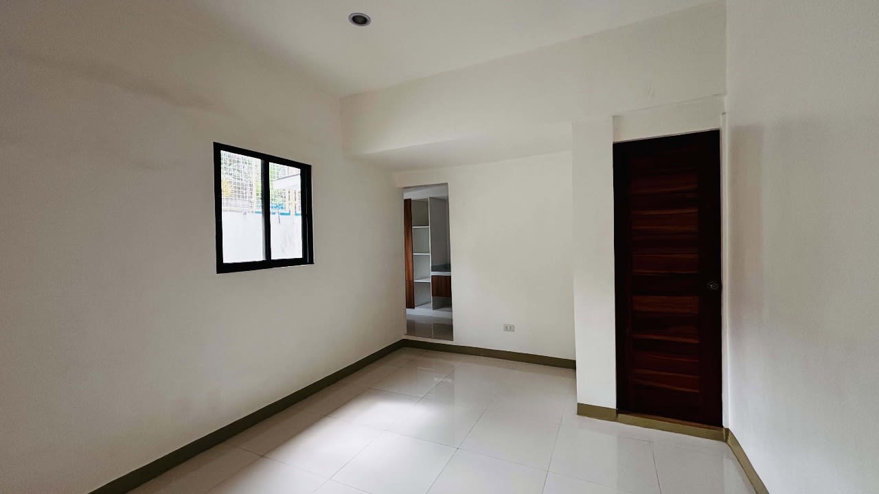 Empty bedroom with white walls, a black-framed window, tiled flooring, and a wooden door in a renovated house for sale in Yati, Liloan.