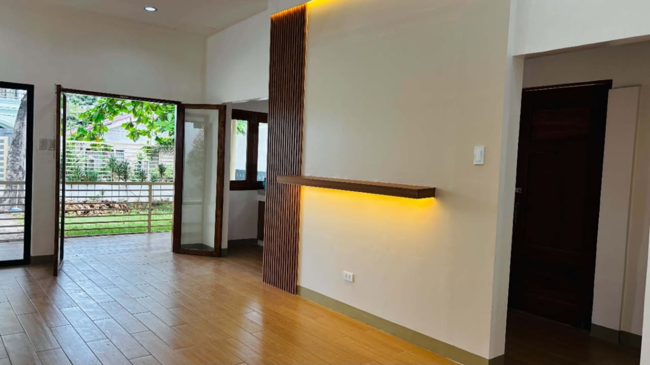 Open-plan living area with wooden flooring, illuminated accent wall, and sliding glass doors leading to the outdoor lawn in Liloan, Cebu.