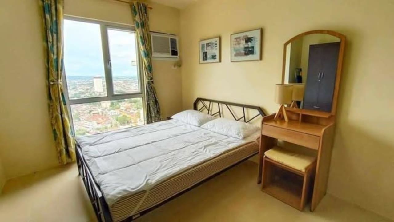 Bed beside a large window with curtains and wooden dressing table in a fully furnished studio condo for sale in Cebu IT Park.