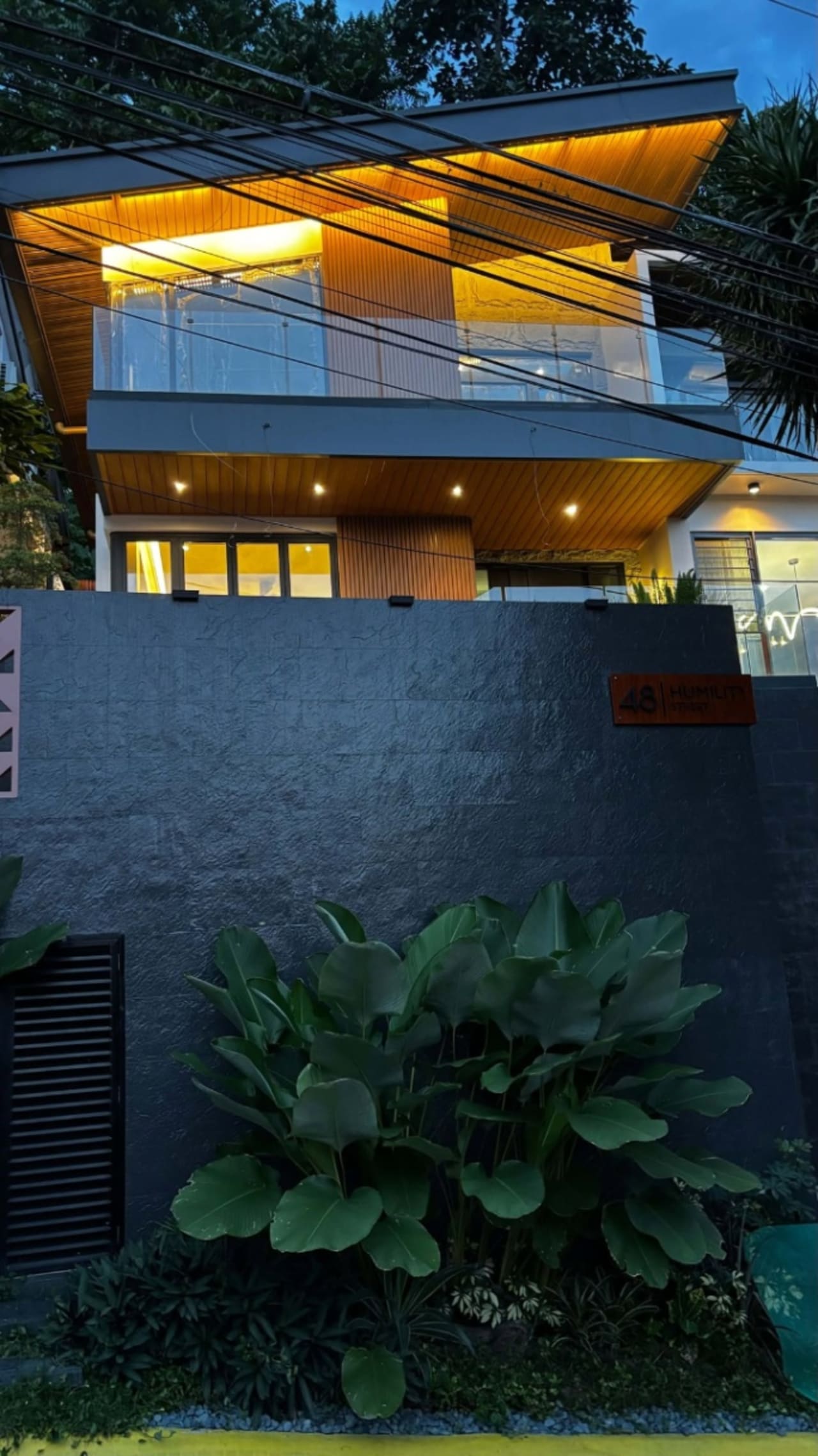 Modern house exterior with a high concrete perimeter wall, illuminated upper-level balcony, glass railing, and landscaped tropical plants in Cebu City.