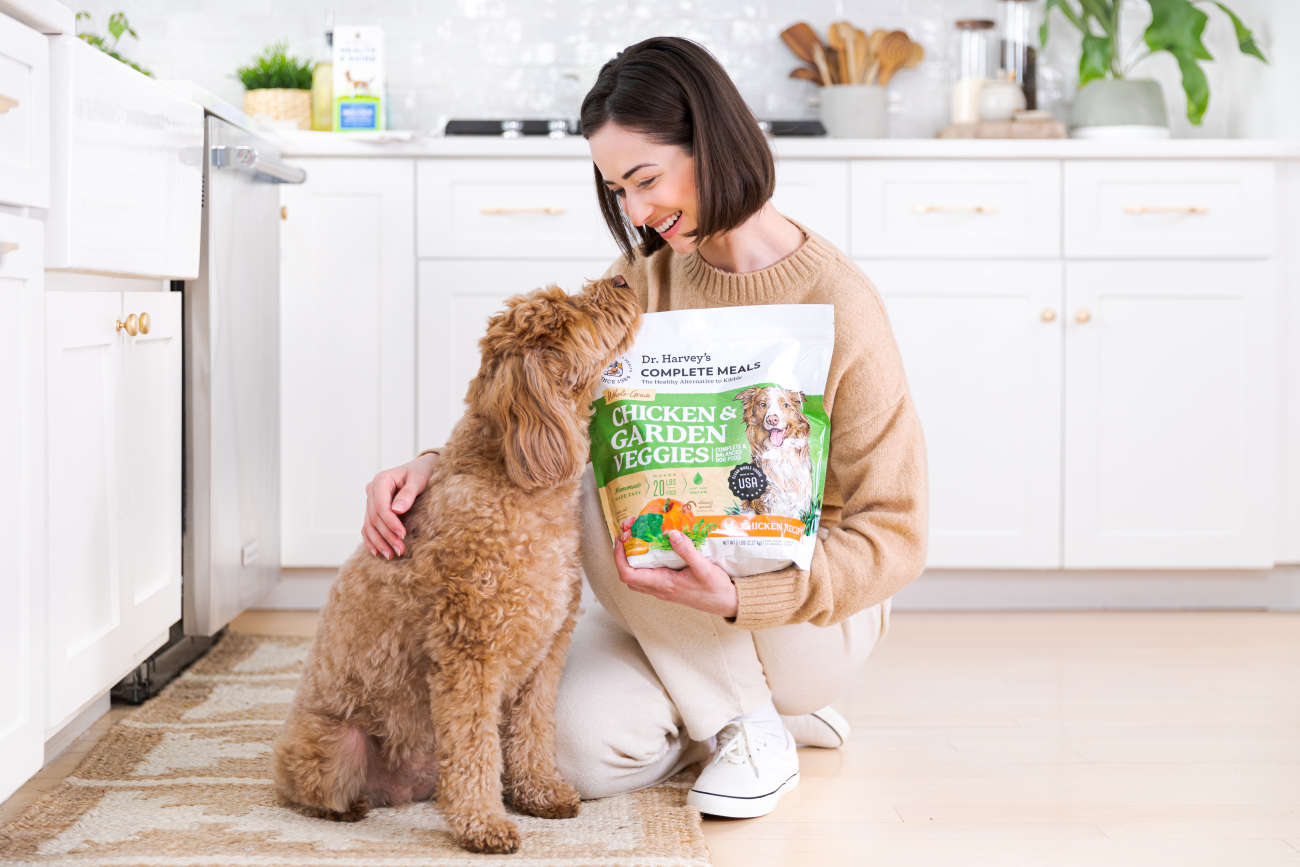 Woman kneeling in a kitchen, holding Dr. Harvey's Chicken & Garden Veggies dog food while her dog sniffs her face