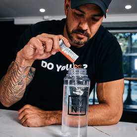 Mind Pump's Adam Schafer adding a Watermelon electrolyte packet to his clear water bottle while resting his left arm on a table.