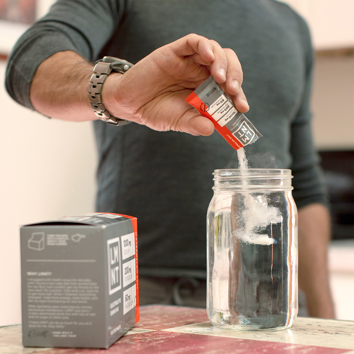 Mind Pump's Adam Schafer adding a Watermelon electrolyte packet to his clear water bottle while resting his left arm on a table.