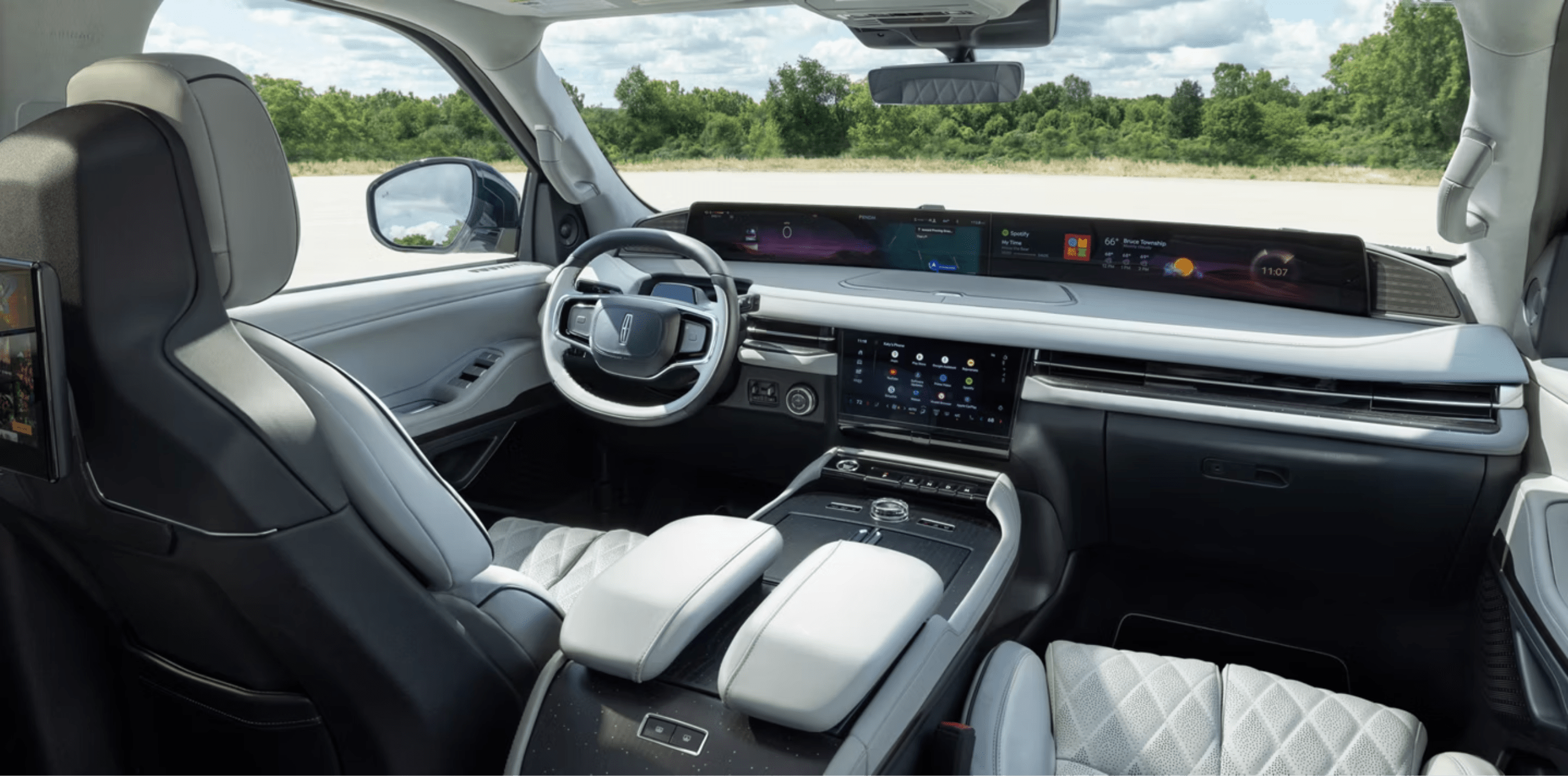 Luxurious interior of a modern car with white and black leather seats and a large touchscreen display, against a backdrop of a bright, overcast sky and a distant tree line.