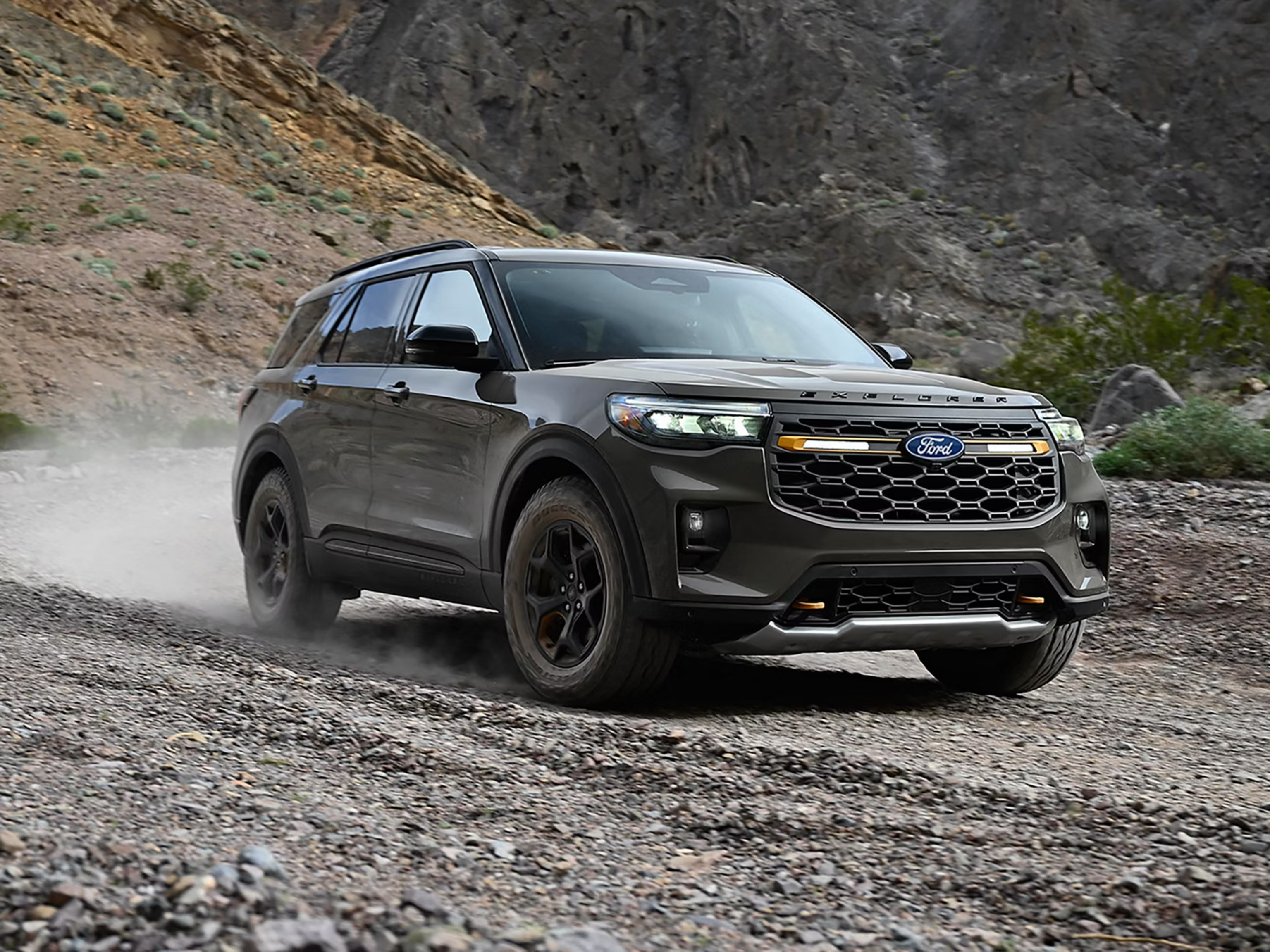 A grey SUV drives across a rocky terrain with a rugged mountain landscape in the background.