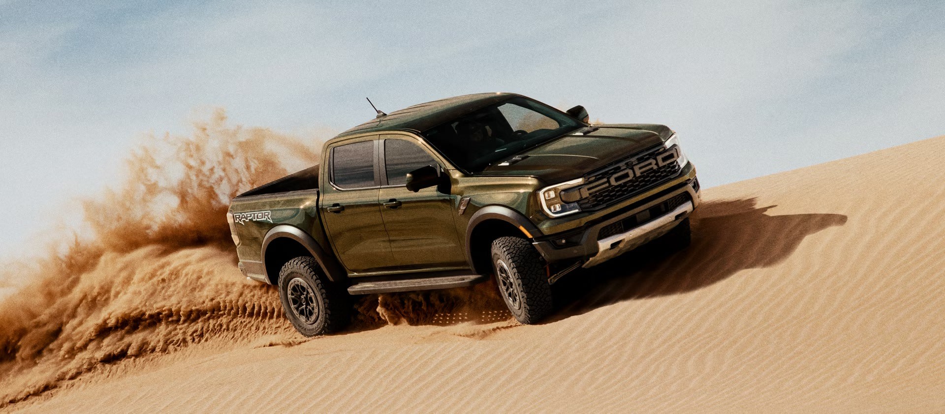 A dark green truck kicks up a large cloud of sand as it drives over a sand dune under a clear blue sky.