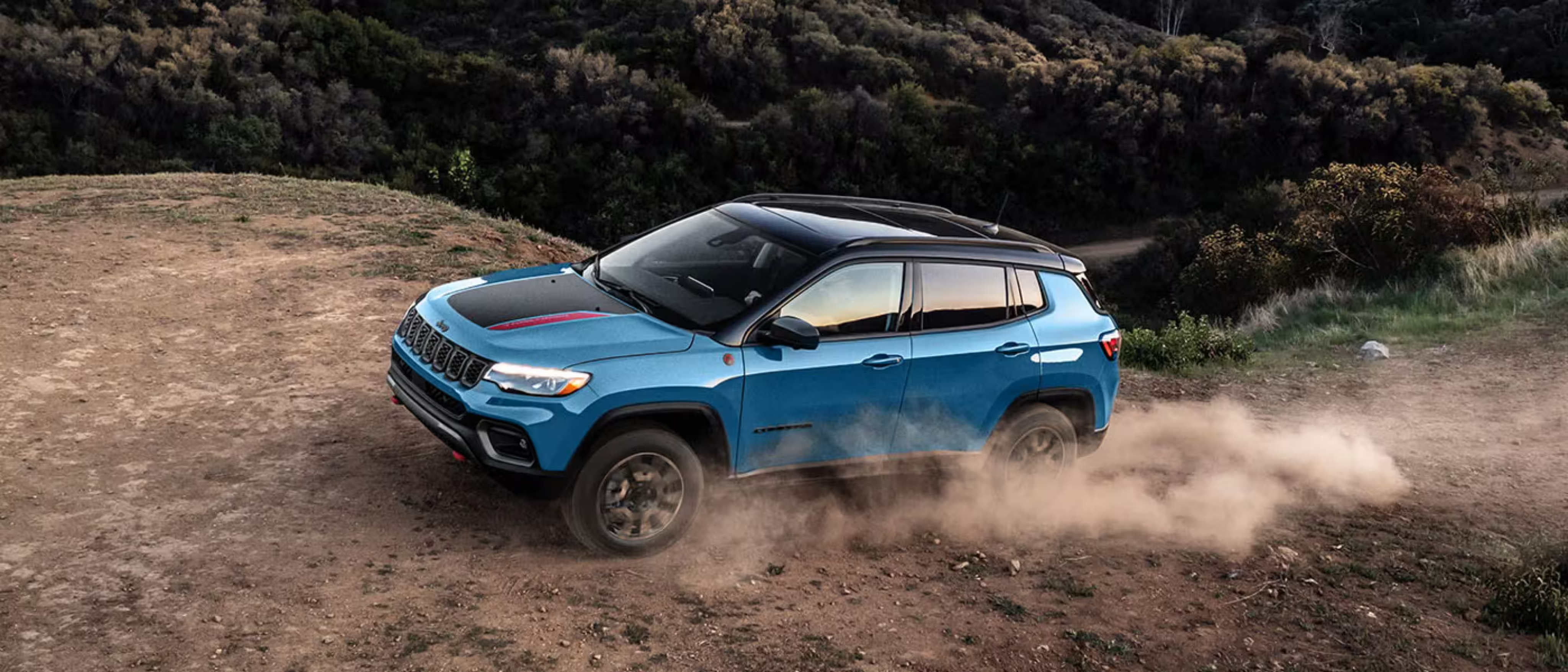 A blue SUV kicks up dust while driving across a dirt path against a backdrop of dense, dark green shrubbery.