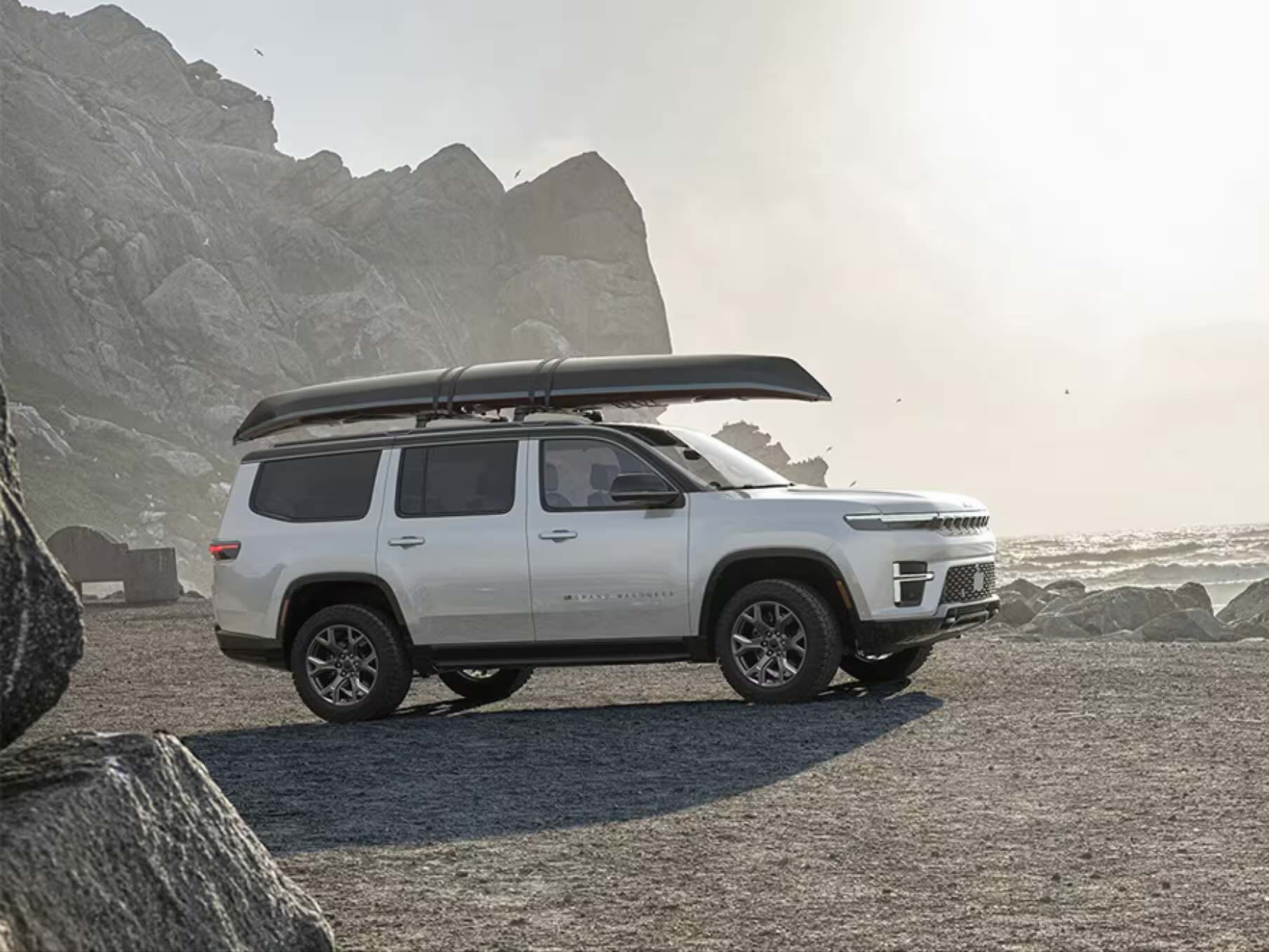 White SUV with a canoe on its roof parked on a rocky shore with ocean and cliffs in the background.
