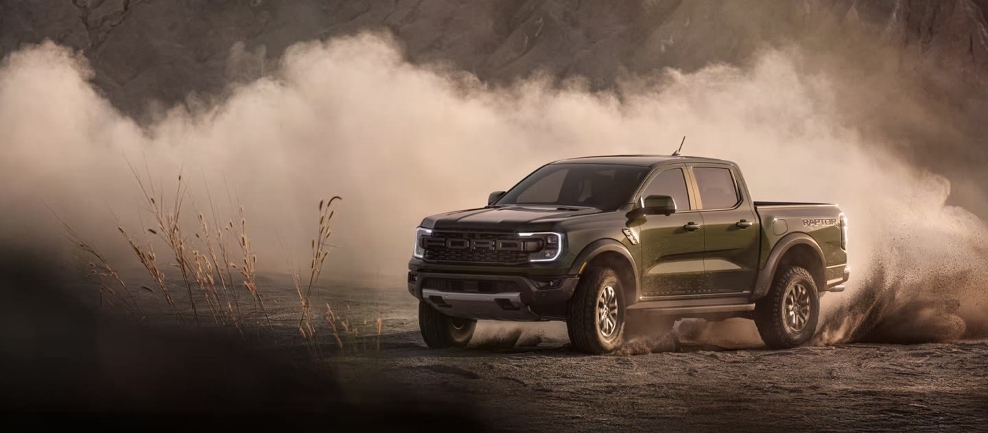 A dark green pickup truck kicks up a cloud of dust as it drives through a barren, rocky landscape.