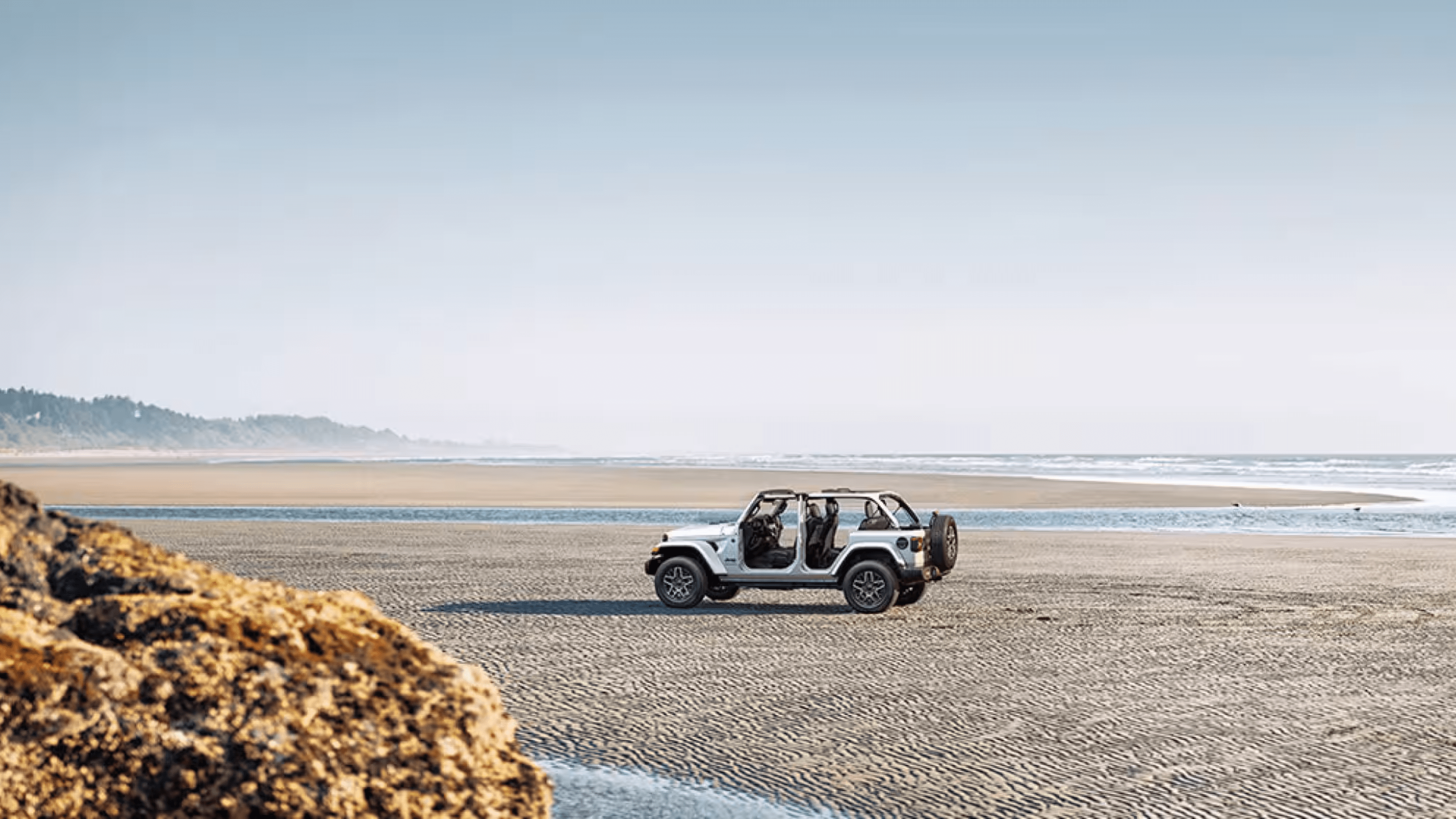 A white SUV parked on a sandy beach with a rocky outcrop in the foreground and a distant coastline under a clear sky.