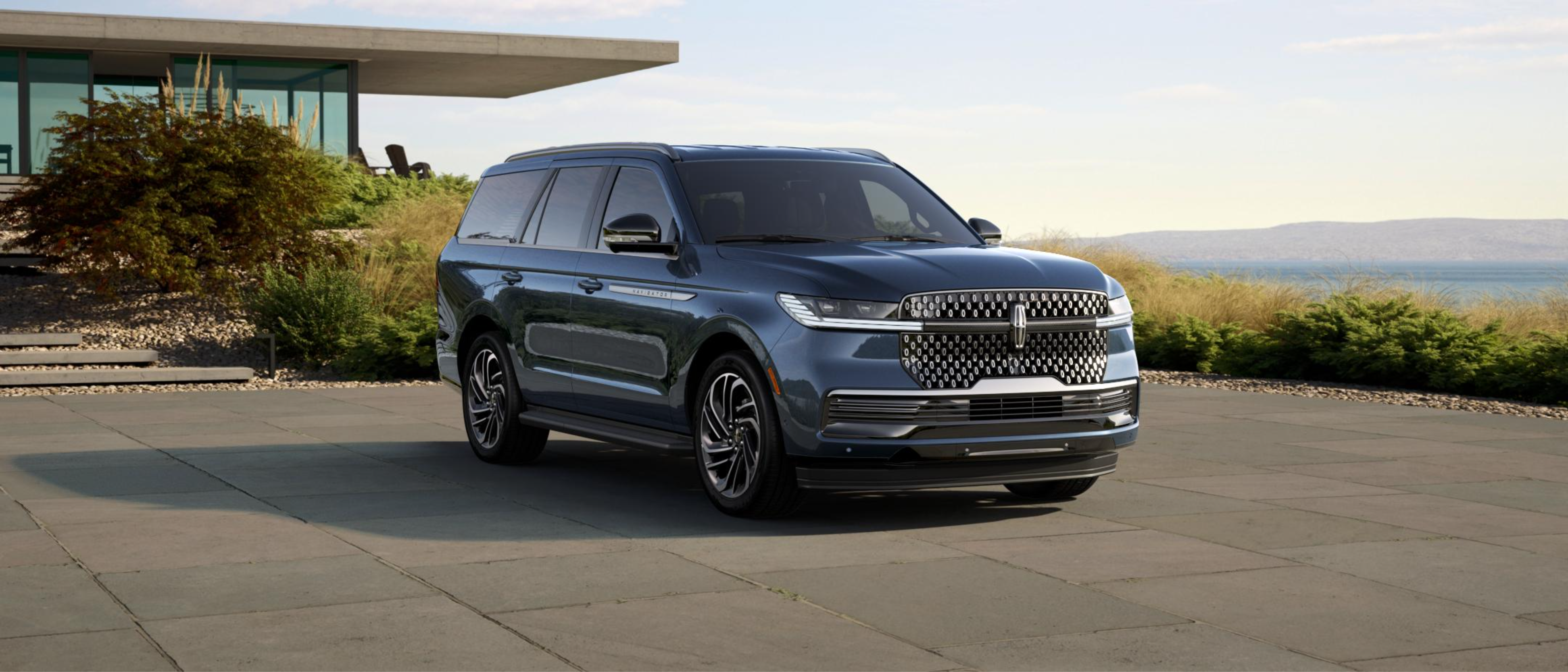Dark blue SUV parked on a paved driveway in front of a modern house with a body of water and hills in the background.