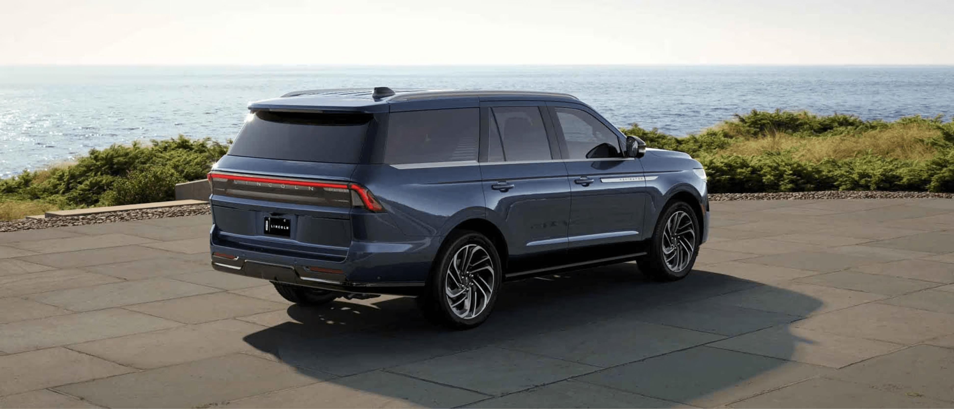 An SUV in blue color parked on a paved area with a scenic view of the ocean and greenery in the background.